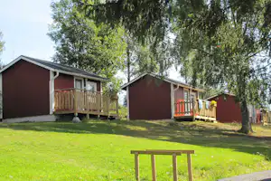 Zwei Camping-Bungalows mit Holzveranda stehen auf einer grünen Wiese, im Vordergrund eine Bank und ein Weg.