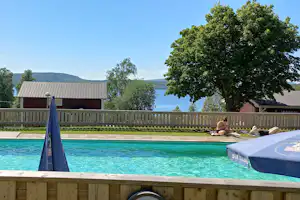 Privater Pool mit Holzumrandung, Liegen und Sonnenschirm vor einem Zaun, dahinter Bäume und See in der Ferne.