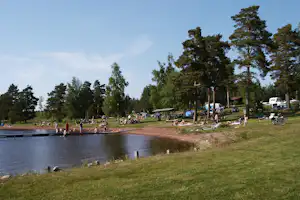 Campingplatz am See mit Grasstrand, Bäumen und Stellplätzen am Ufer, Boote und Wohnwagen im Hintergrund.