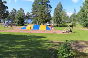 Spielplatz auf dem Campingplatz mit gelbem und blauem Hüpfbereich, Rutsche und Spielgeräten auf Rasen zwischen Bäumen.