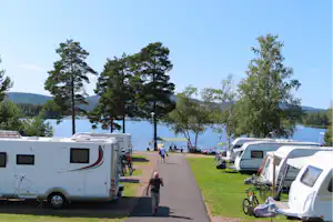 Wohnmobile auf einem Campingplatz am See, mit Rasenflächen, Stellplätzen, Fahrrädern und einem Weg zwischen den Fahrzeugen.