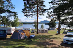 Zelt auf einem Campingplatz am See, mit Wohnmobil, Auto und weiteren Stellplätzen zwischen hohen Bäumen.