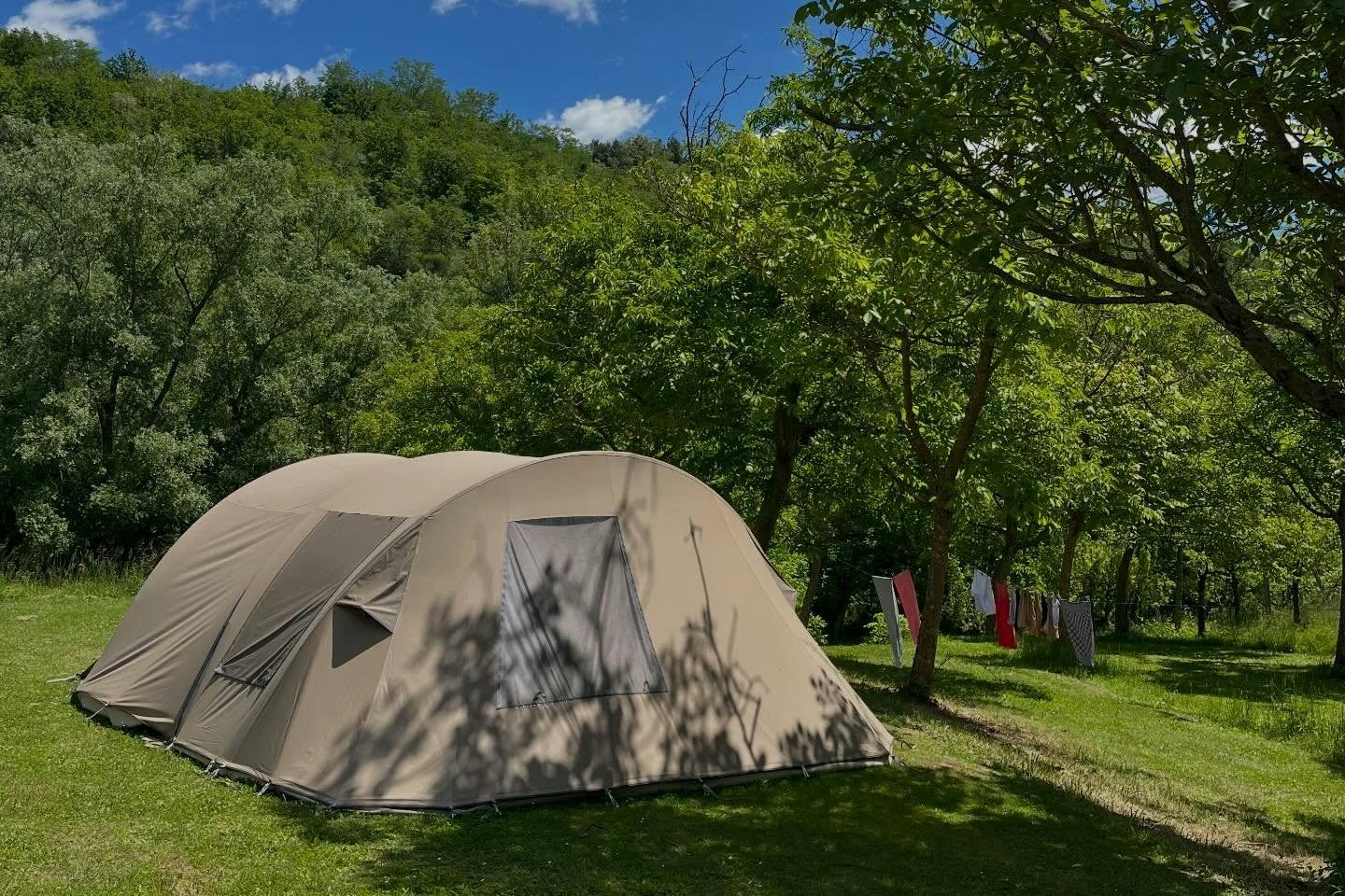 Zelt auf einem grünen Campingplatz unter Bäumen, mit Grasweg und blauem Himmel mit Wolken im Hintergrund.