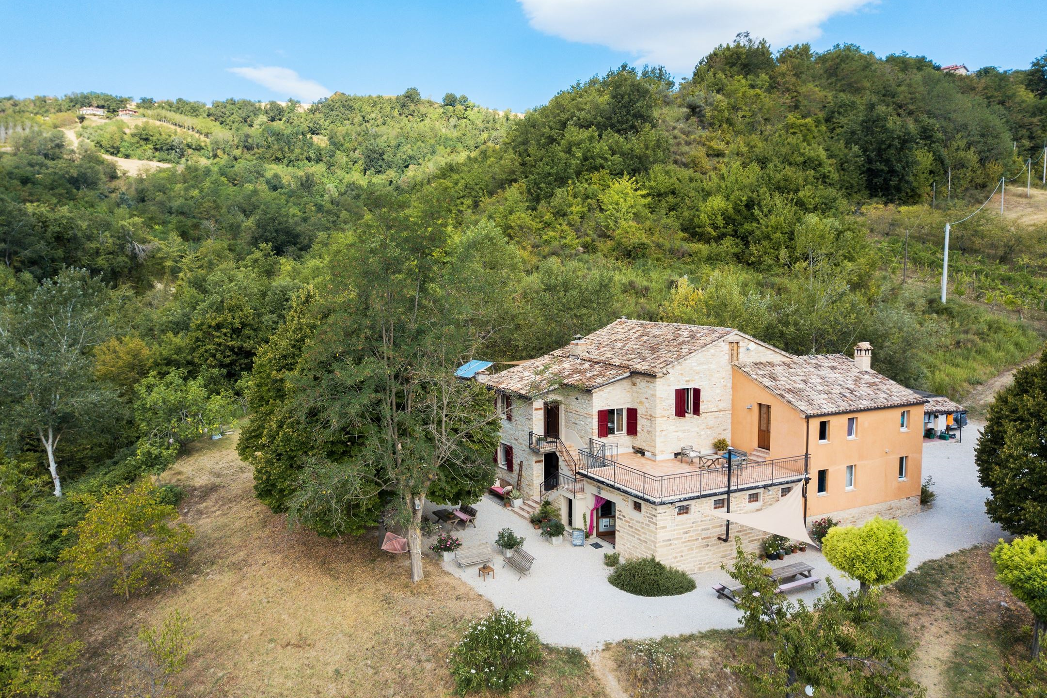 Ferienhaus mit mehreren Etagen und roten Fensterläden, Terrasse und Garten am Hang, umgeben von Bäumen und grünen Hügeln.