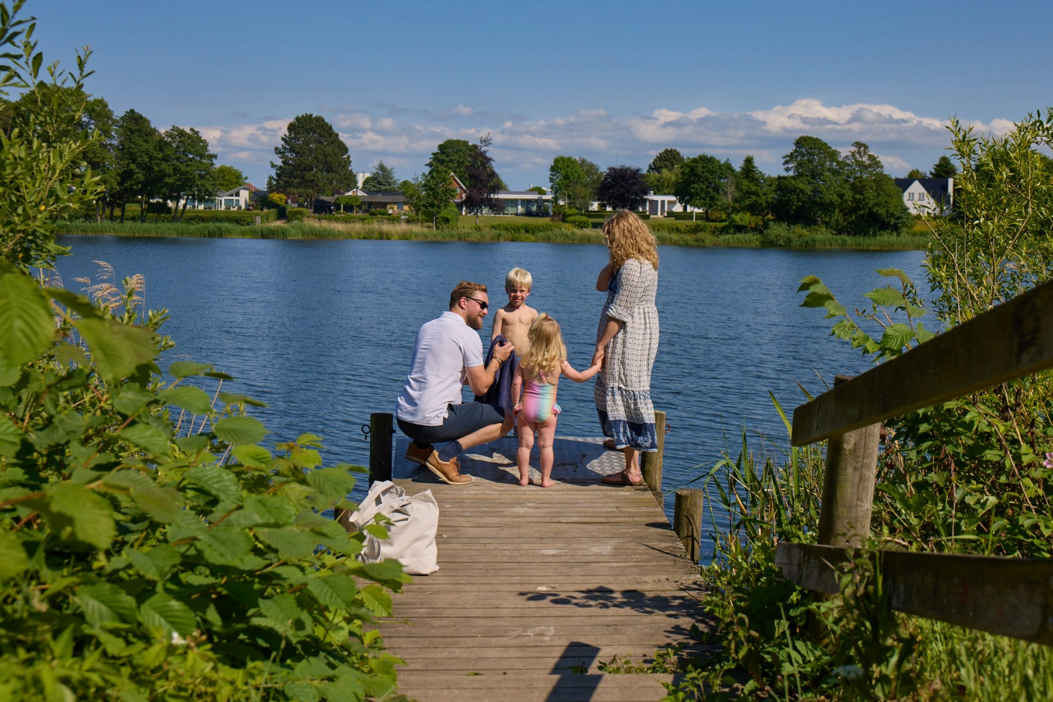 Familie sitzt auf einer Holzplattform am See, im Hintergrund Bäume und Häuser am Ufer.