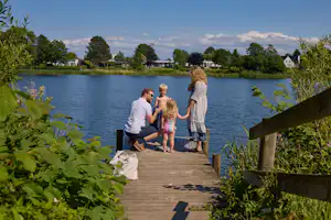 Familie sitzt auf einer Holzplattform am See, im Hintergrund Bäume und Häuser am Ufer.