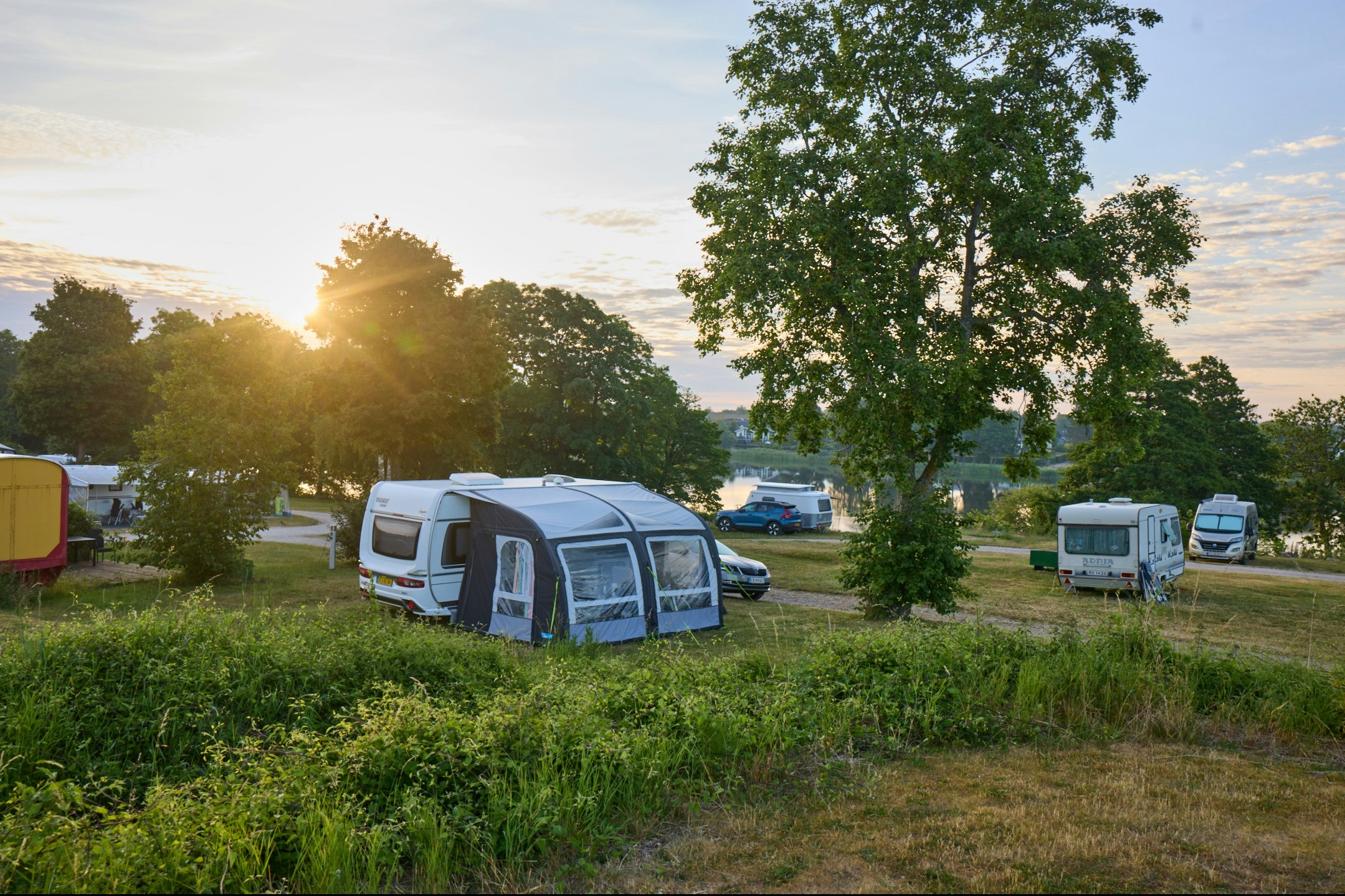Campingplatz mit Wohnwagen und Vorzelten, daneben geparkte Autos, im Hintergrund Bäume und ein Weg.
