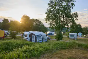 Campingplatz mit Wohnwagen und Vorzelten, daneben geparkte Autos, im Hintergrund Bäume und ein Weg.