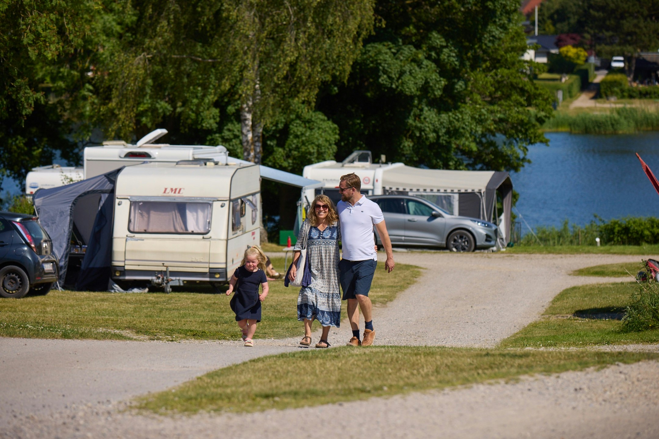 Familie geht auf dem Campingplatz entlang des Weges, neben Wohnmobilen und einem Zelt, im Hintergrund Bäume und Autos.