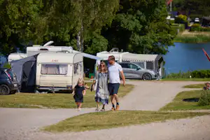 Familie geht auf dem Campingplatz entlang des Weges, neben Wohnmobilen und einem Zelt, im Hintergrund Bäume und Autos.