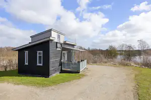 Moderner Camping-Bungalow mit Terrasse und Glasgeländer, neben einem Kiesweg, umgeben von Rasen und Bäumen.