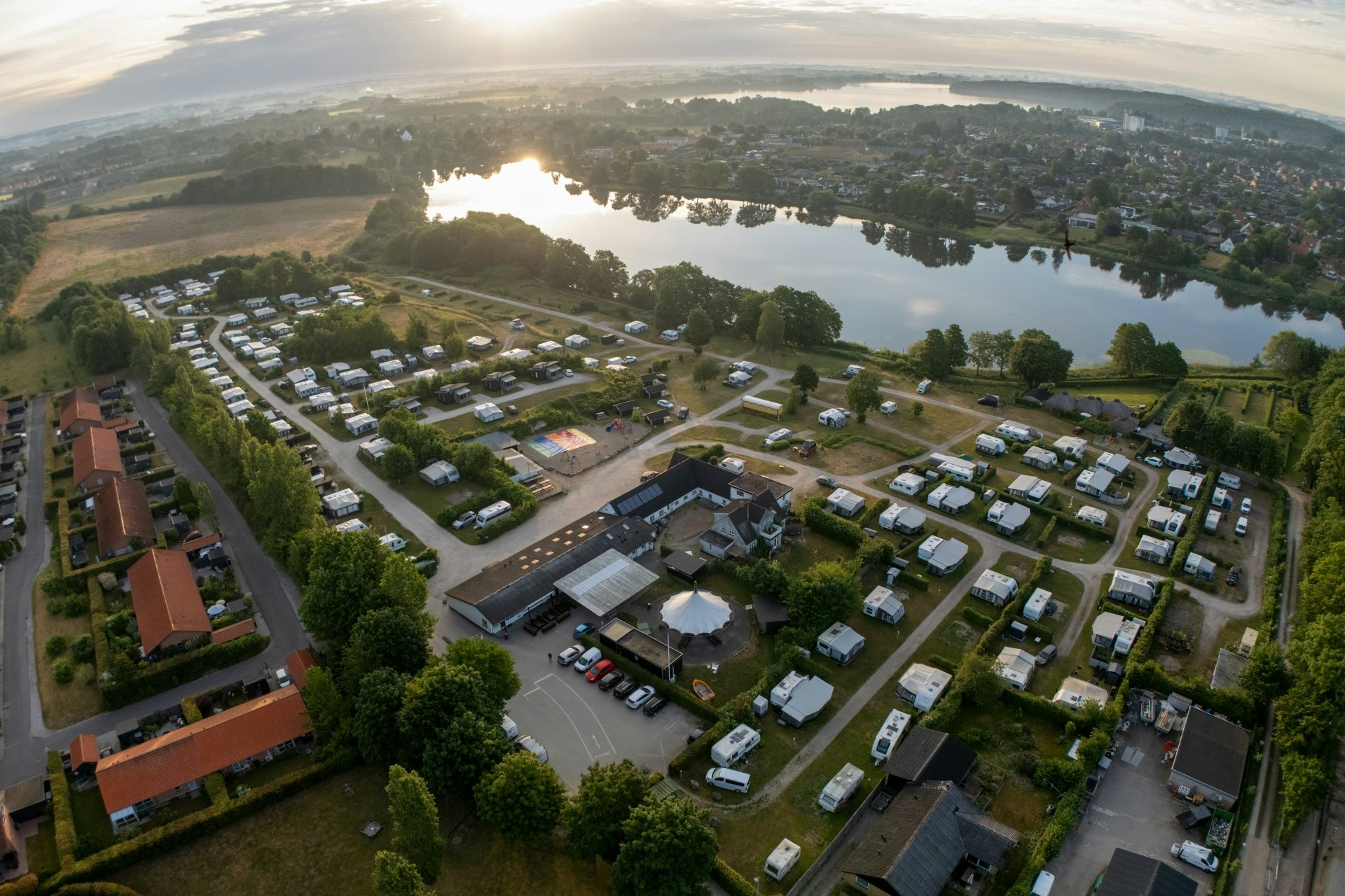 Luftaufnahme eines Campingplatzes mit vielen Stellplätzen, Wohnwagen und Zelten, Wegen, Bäumen und einem großen See im Hintergrund.