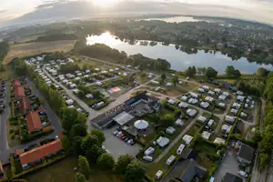 Luftaufnahme eines Campingplatzes mit vielen Stellplätzen, Wohnwagen und Zelten, Wegen, Bäumen und einem großen See im Hintergrund.