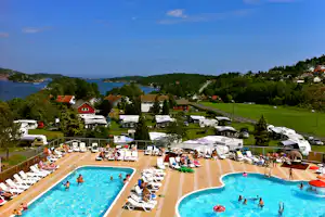 Blick auf den Campingplatz mit Pool, Liegestühlen und Wohnwagen, dahinter grünes Ufer und See.