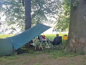Campingzelt am Waldrand mit zwei Campingstühlen und Tisch, neben einem Baum und unter grünen Blättern.