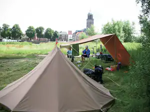 Campingzelt im Vordergrund, daneben weitere Zelte auf einer Wiese; im Hintergrund ein Kirchturm und Bäume.