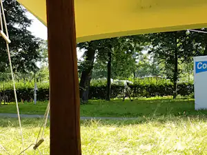 Grüne Campinganlage mit Rasen, Hecken und Bäumen, im Vordergrund ein gelbes Zeltgestänge und ein Schild.
