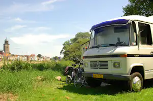 Weißes Wohnmobil auf grünem Gras am Campingplatz, daneben ein Fahrrad, im Hintergrund Bäume und eine Stadt mit Kirchturm.