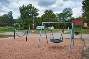 Spielplatz am Campingplatz mit blauen Schaukeln, Sandboden, Bäumen und einem roten Dachgebäude im Hintergrund.