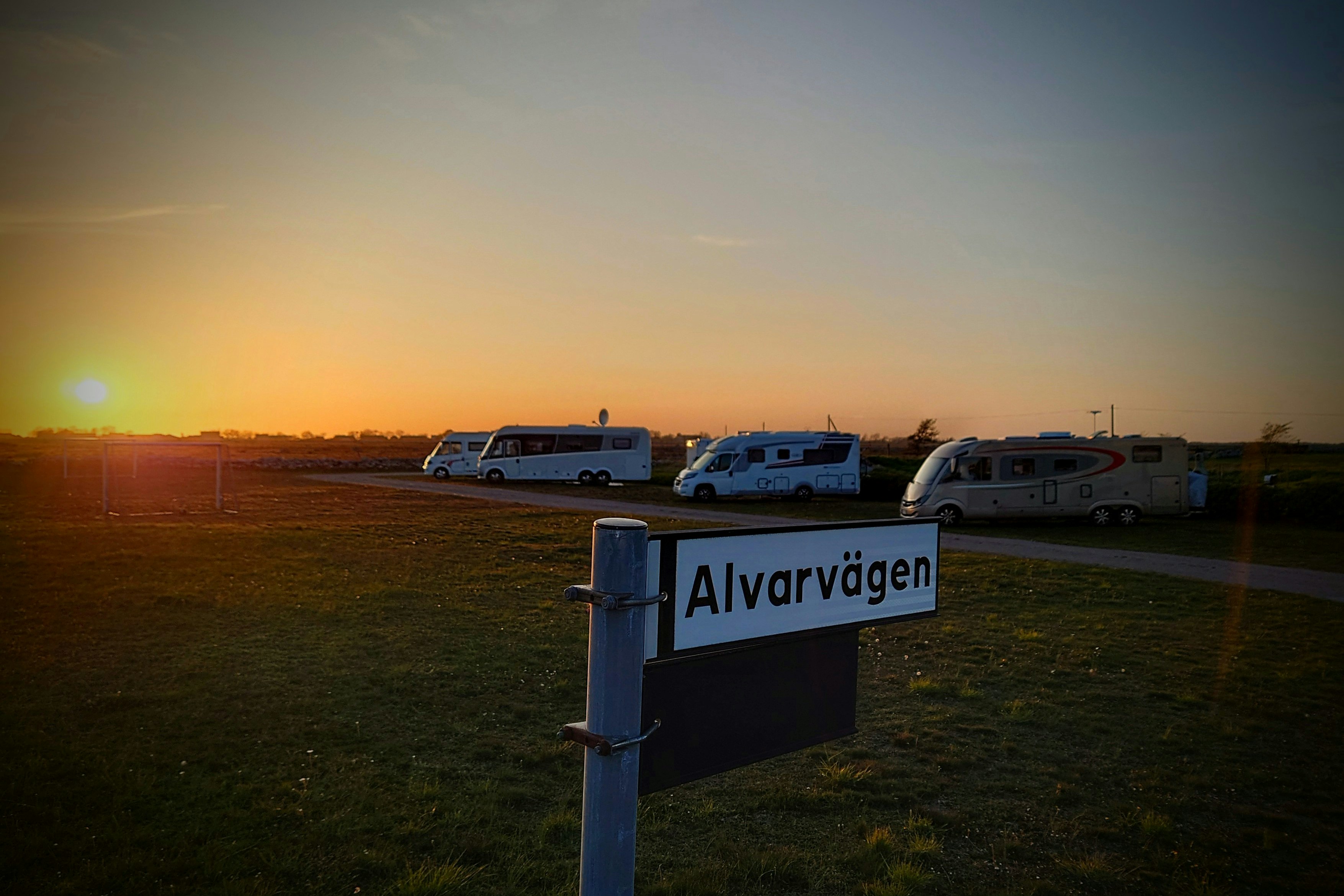 Schild „Alvarvagen“ am Campingplatz, im Hintergrund Wohnwagen und Wohnmobile auf einer Wiese bei Sonnenuntergang.