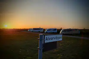 Schild „Alvarvagen“ am Campingplatz, im Hintergrund Wohnwagen und Wohnmobile auf einer Wiese bei Sonnenuntergang.