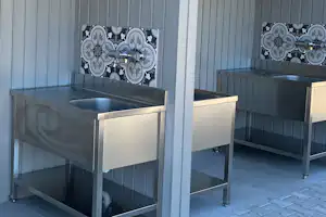Überdachter Küchenbereich mit zwei Spülen und Arbeitsplatten, daneben Stühle und Fliesenboden am Campingplatz.