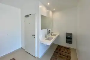 Modernes Badezimmer mit weißem Waschbecken, Spiegel, Handtuchhalter und Tür; Fliesenboden und Wandheizung im Hintergrund.