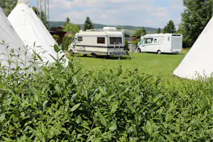 Zelteingang im Vordergrund, dahinter Wohnwagen und Wohnmobil auf einer grünen Campingwiese mit Bäumen und Hügeln.