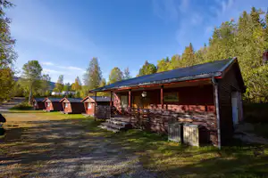 Holzhütte mit Veranda und Stufen, daneben weitere Campinghütten im Grünen, Bäume und Rasenfläche am Weg.