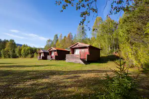 Rote Holzhütten auf einem Campingplatz im Grünen, umgeben von Bäumen und Rasenfläche.