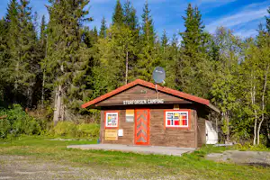 Holzhütte mit roter Tür und Beschriftung „STURFURSEN CAMPING“, davor Rasen und Weg, umgeben von hohen Nadelbäumen.