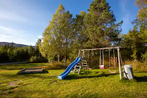 Spielplatz am Campingplatz: Schaukelgestell mit Rutsche, Sandkasten und Mülleimer auf einer grünen Wiese, umgeben von Bäumen.