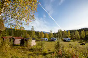 Campingwiese mit mehreren Wohnwagen, kleinen Gebäuden und Bäumen, im Hintergrund Hügel und Berge.