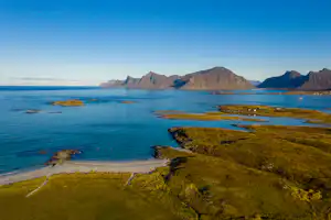 Blaue Bucht mit Felseninseln und Küstenlinie, grünes Ufer mit Wegen, dahinter hohe Berge im klaren Himmel.