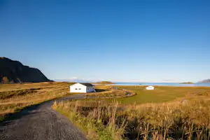 Weiße Camping-Häuser auf einer Wiese nahe dem Meer, mit Schotterweg und Grasbüscheln im Vordergrund.