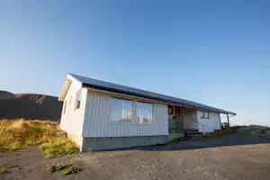 Weißes Campinghaus mit Fensterfront und überdachtem Eingang, am Hang neben einem Kiesweg und Gras.