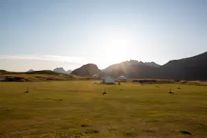 Campingwiese mit zwei Wohnwagen auf einer grünen Fläche, im Hintergrund Berge und tief stehende Sonne am Himmel.