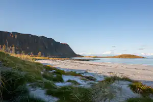 Sandstrand mit Dünen und Gras vor der Küste, im Hintergrund Klippen und das ruhige Meer.