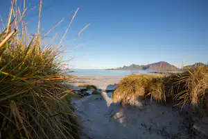 Sanddünen mit Gras im Vordergrund, dahinter das Meer und entfernte Berge unter klarem Himmel.