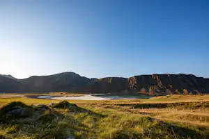 Blick auf eine weite Landschaft mit See, Wiesen und Bergen im Hintergrund, umgeben von Felsen und Gras.