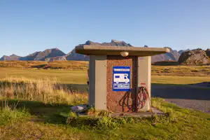 Camping-Anschlusskasten aus Stein mit Kabeltrommel, auf einer Wiese vor Bergen und weitem Feld.