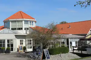 Campinghaus mit rotem Ziegeldach, Terrasse und großen Fenstern; davor Fahrräder und ein gepflasterter Weg.