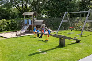 Spielplatz mit Holz-Klettergerüst, Rutsche, Schaukeln und Federwippe auf grünem Rasen, umgeben von Hecken und Bäumen.