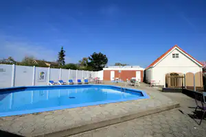 Blauer Campingpool mit Leiter, Sonnenliegen und Tischen vor weißen Gebäuden und Zäunen, im Hintergrund Bäume und Himmel.