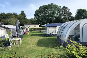 Campingzelte auf einem Rasenplatz, mit Gartenmöbeln und einem Wohnwagen im Hintergrund, umgeben von Hecken und Bäumen.