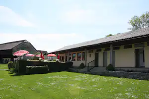 Campinggebäude mit Terrasse, Stufen und Rasenfläche, davor rote Sonnenschirme und Hecken im Garten.