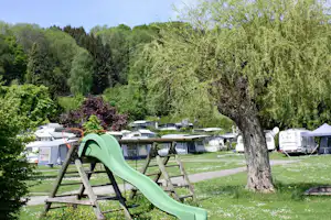 Grüne Rutsche auf einem Spielplatz, im Hintergrund Wohnwagen und Zelte auf dem Campingplatz, umgeben von Bäumen und Rasen.