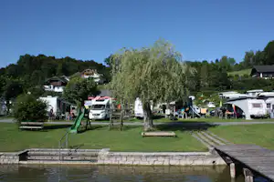 Campingplatz mit Wohnmobilen und Wohnwagen, Sitzgelegenheiten und Zeltplanen unter Bäumen am grünen Weg.