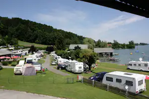 Campingplatz am See mit Wohnmobilen, Zelten und Stellplätzen am Ufer, im Hintergrund Stege und Boote.