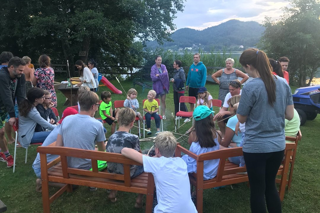 Familie sitzt im Kreis auf Holzstühlen im Campinggarten, im Hintergrund Bäume, See und Berge.